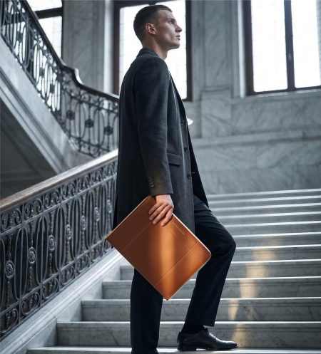 A male model in a high-fashion designer outfit holding the Stefan Serval Entrata Superiore luxury cognac leather portfolio while standing on modern concrete architectural stairs.